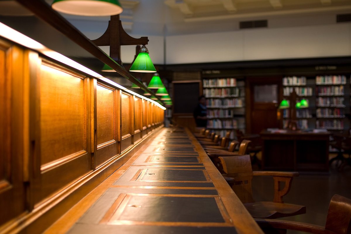 A long wooden library reading desk lit by a row of green glass shaded lamps, with book stacks and wooden chairs behind it.