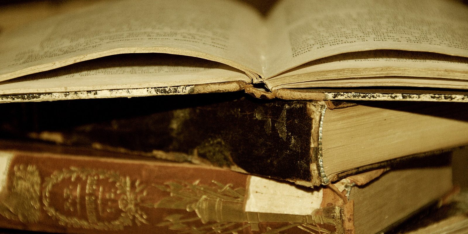 An open book with yellowed pages resting on a stack of worn leather bound volumes, one with a gilt embossed red spine.