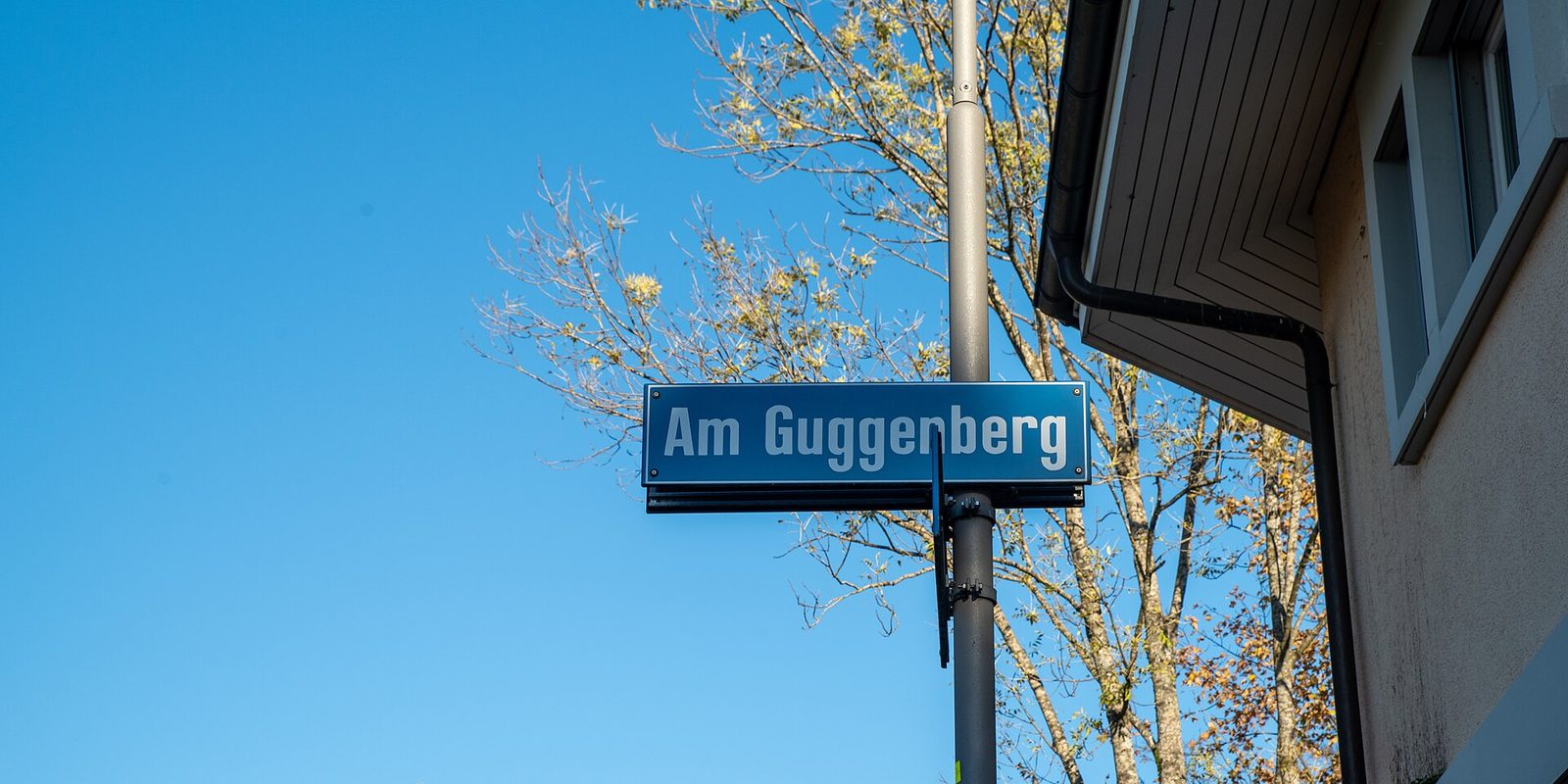 A blue enamel street sign reading Am Guggenberg bolted to a metal pole under clear sky, with a bare tree and a stucco house eave behind.