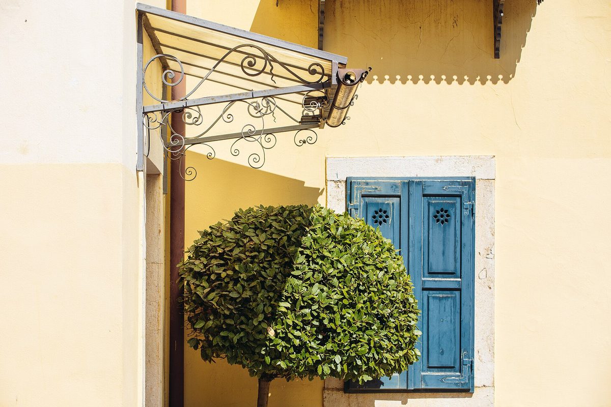 Closed blue louvered wooden shutters on a pale yellow stucco wall, beside a clipped round-topped tree and a scrolled wrought-iron awning frame.