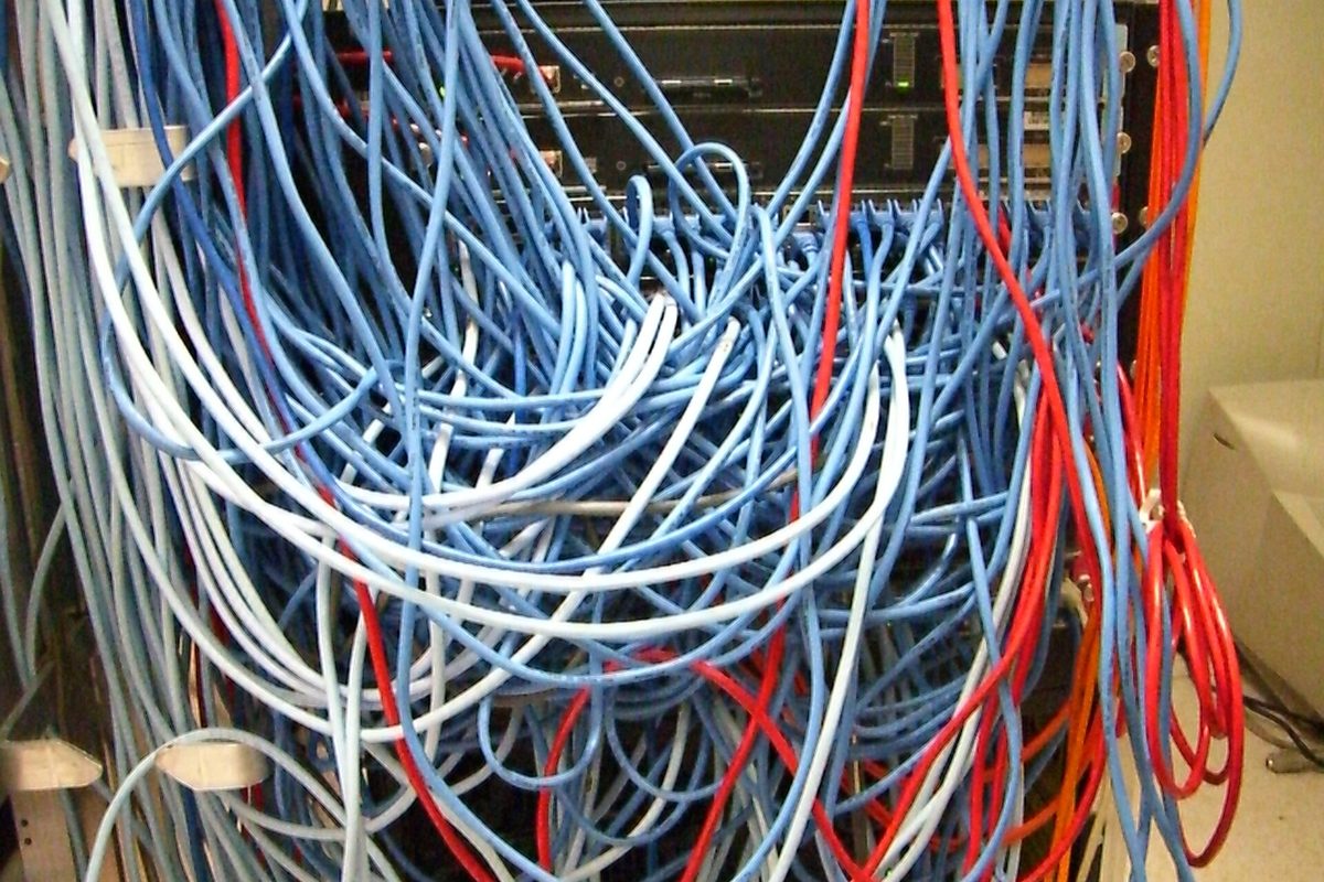 A dense tangle of blue, white and red network cables looping down from a black equipment rack in a wiring closet.