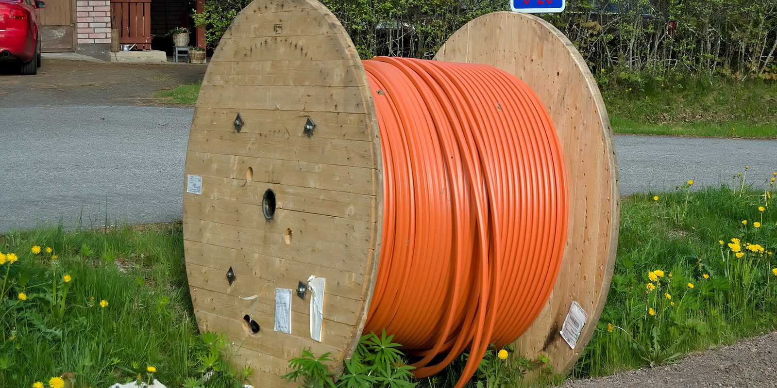 A large wooden cable spool wound with coiled orange conduit, standing in grass and dandelions beside a gravel driveway.