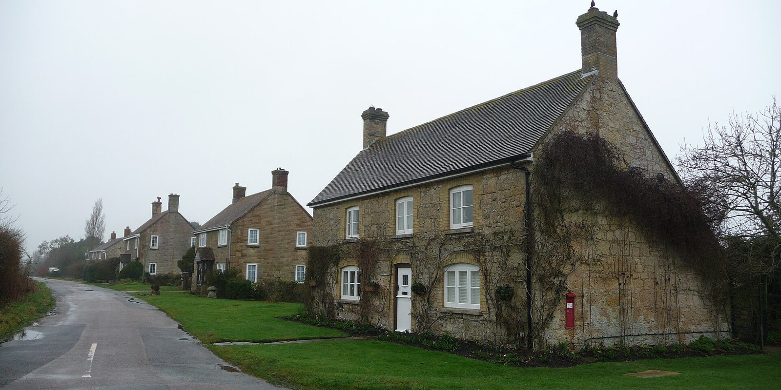 A red postbox set into the gable of a golden stone cottage with a tiled roof and chimneys, along a wet lane of houses under grey mist.