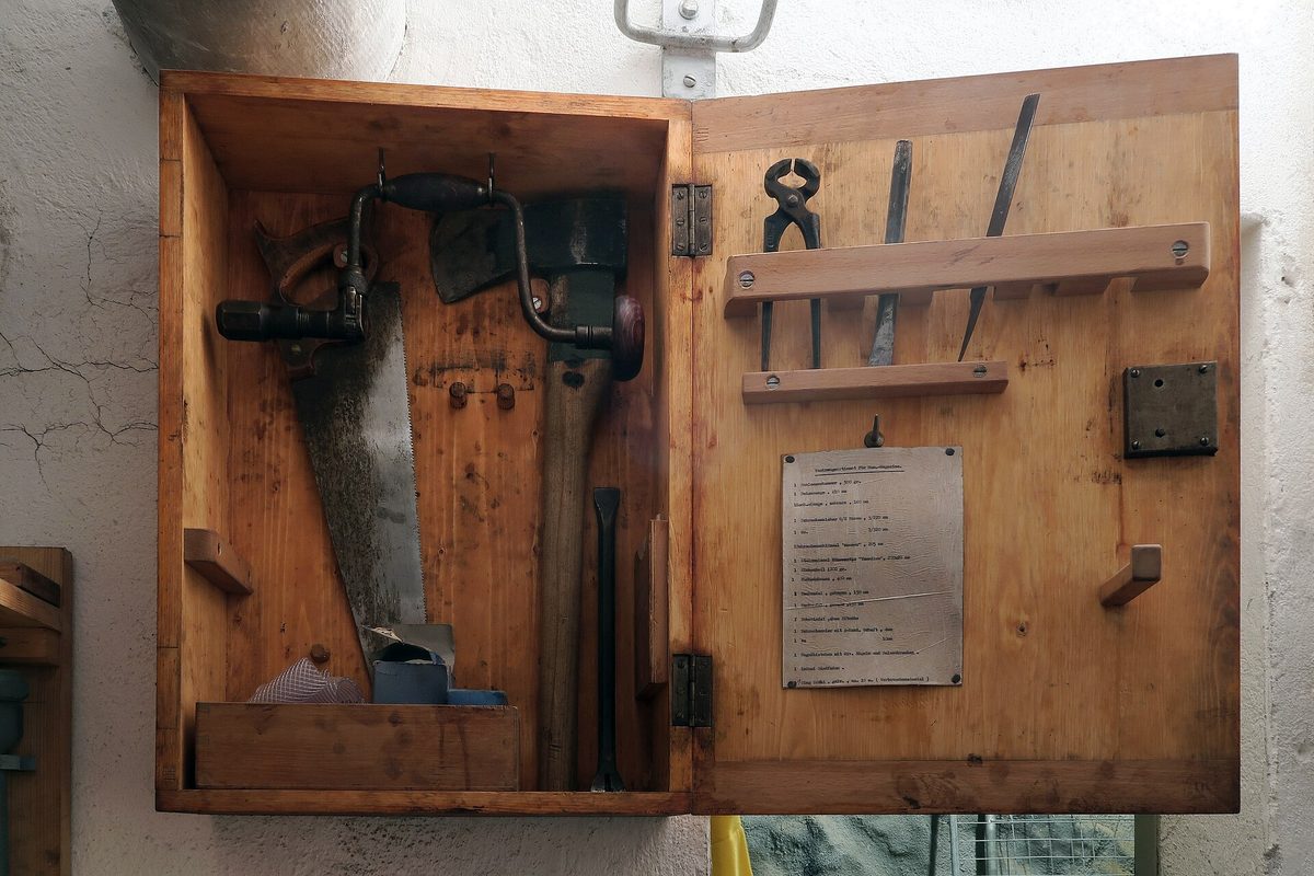 An open wooden wall cabinet holding a hand saw, brace drill, hatchet, chisel, files and pincers, with a typed metal list on the door.