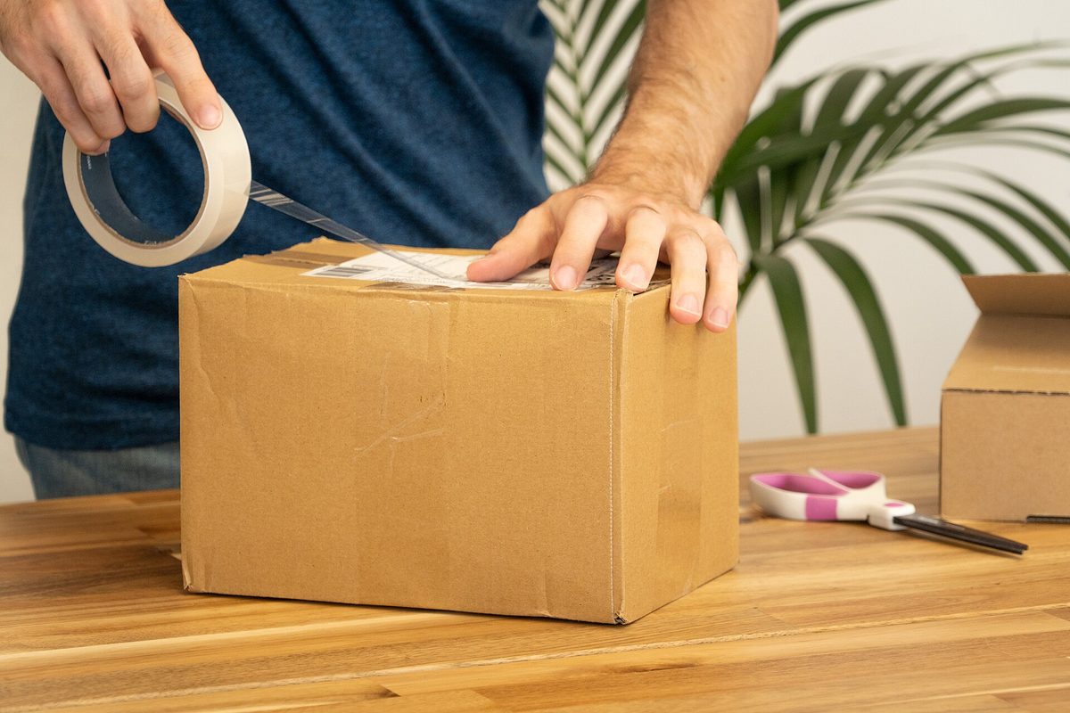 A man in a navy shirt pulling clear tape from a roll across the top of a cardboard box on a wooden table, scissors resting beside it