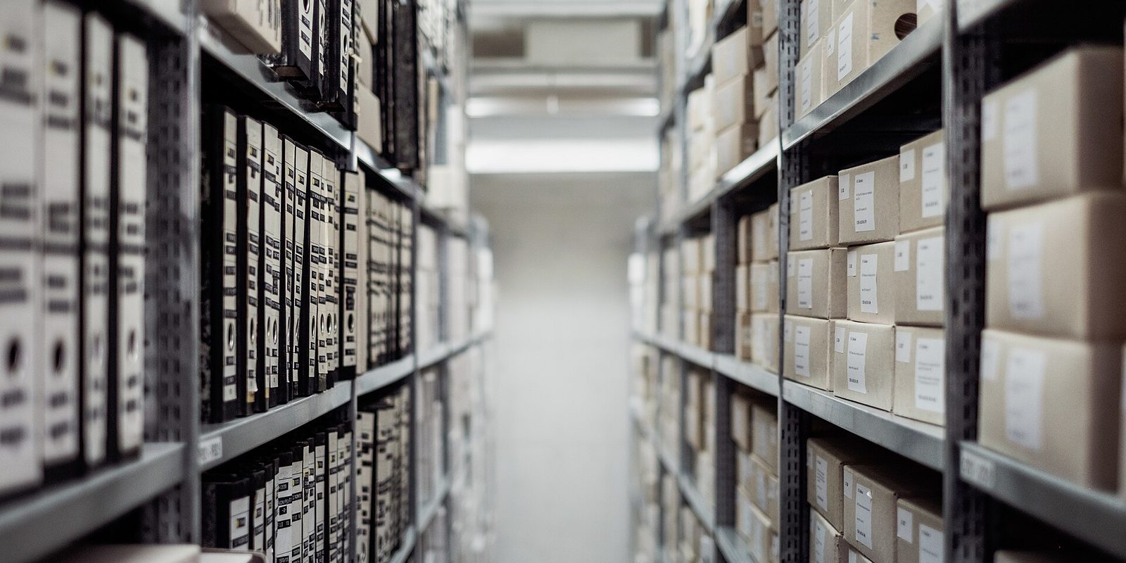 A narrow storage aisle between metal shelving, black and white ring binders on the left and labelled cardboard document boxes on the right.