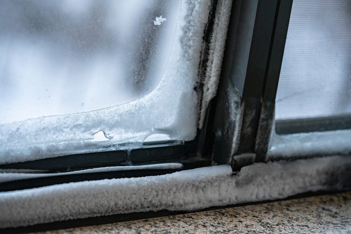 Thick frost and snow built up along the black rubber seal and aluminium frame of a window corner beside a mesh screen.