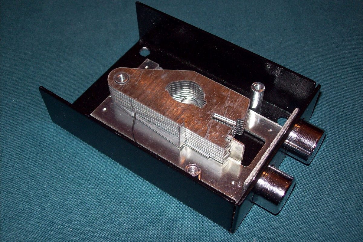 A machined steel lock mechanism with two chrome bolts inside a black metal case, resting on teal fabric.