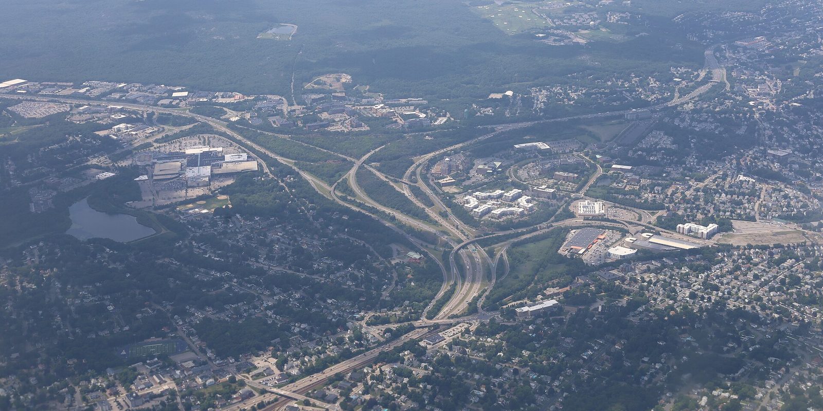Wooded suburban sprawl seen from high above, with a curving highway interchange, low office buildings, a small lake and hazy green hills