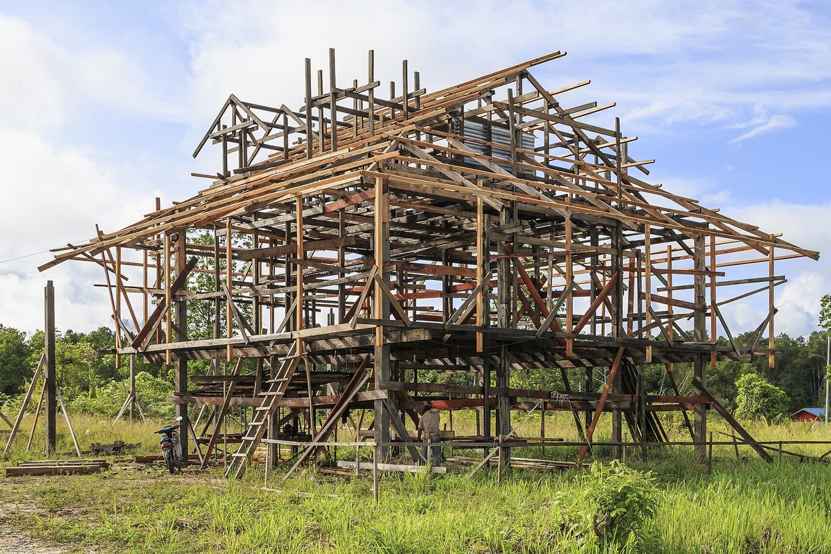 The bare timber frame of a large stilted house under construction, rough wooden beams and rafters against a blue sky, a man working below