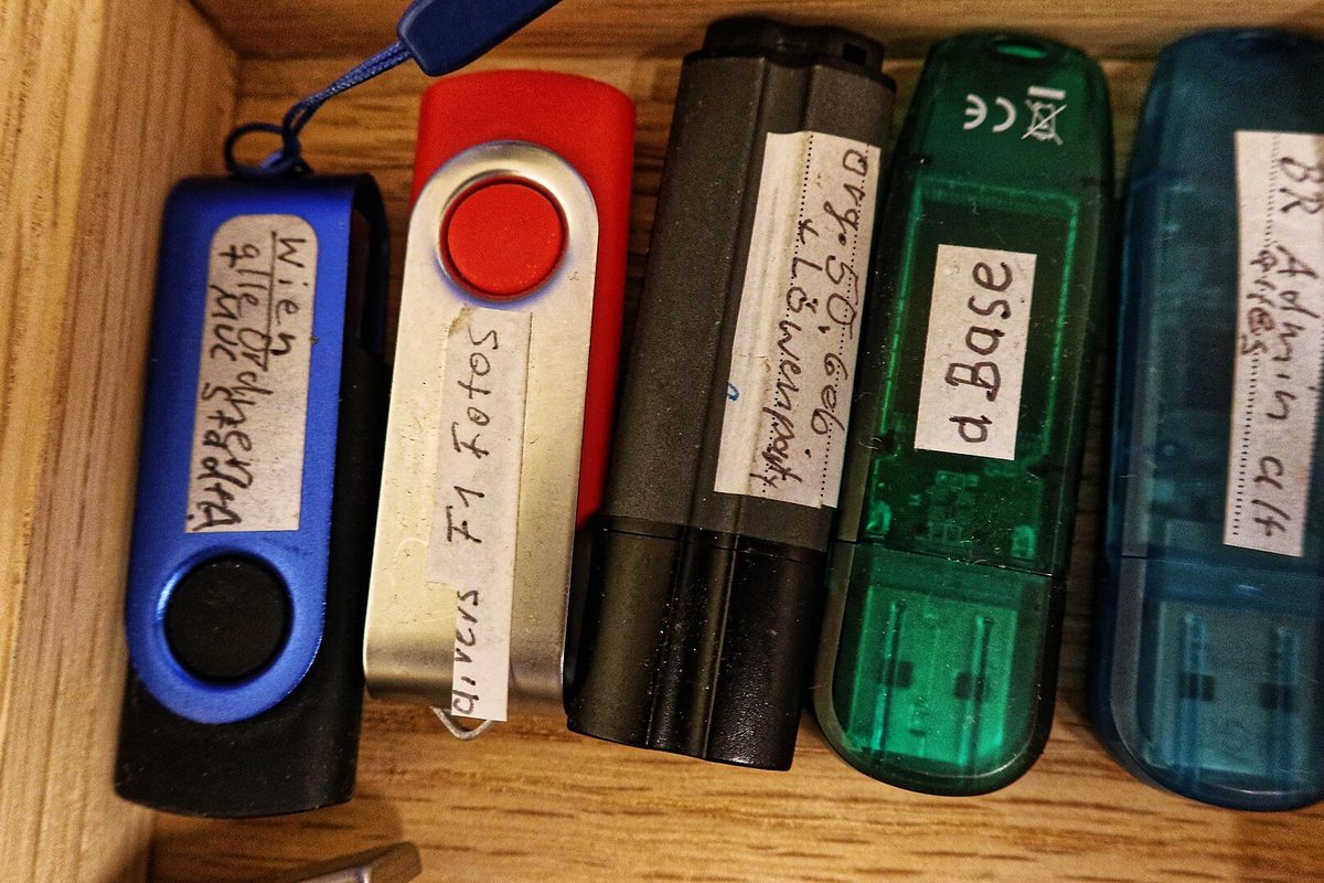 Five USB flash drives in blue, red, black, green and teal lined up in a wooden drawer, each wearing a handwritten tape label
