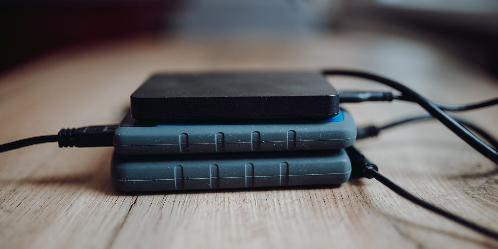 Three portable external hard drives stacked on a wooden tabletop, black and grey plastic cases with USB cables trailing off to the side