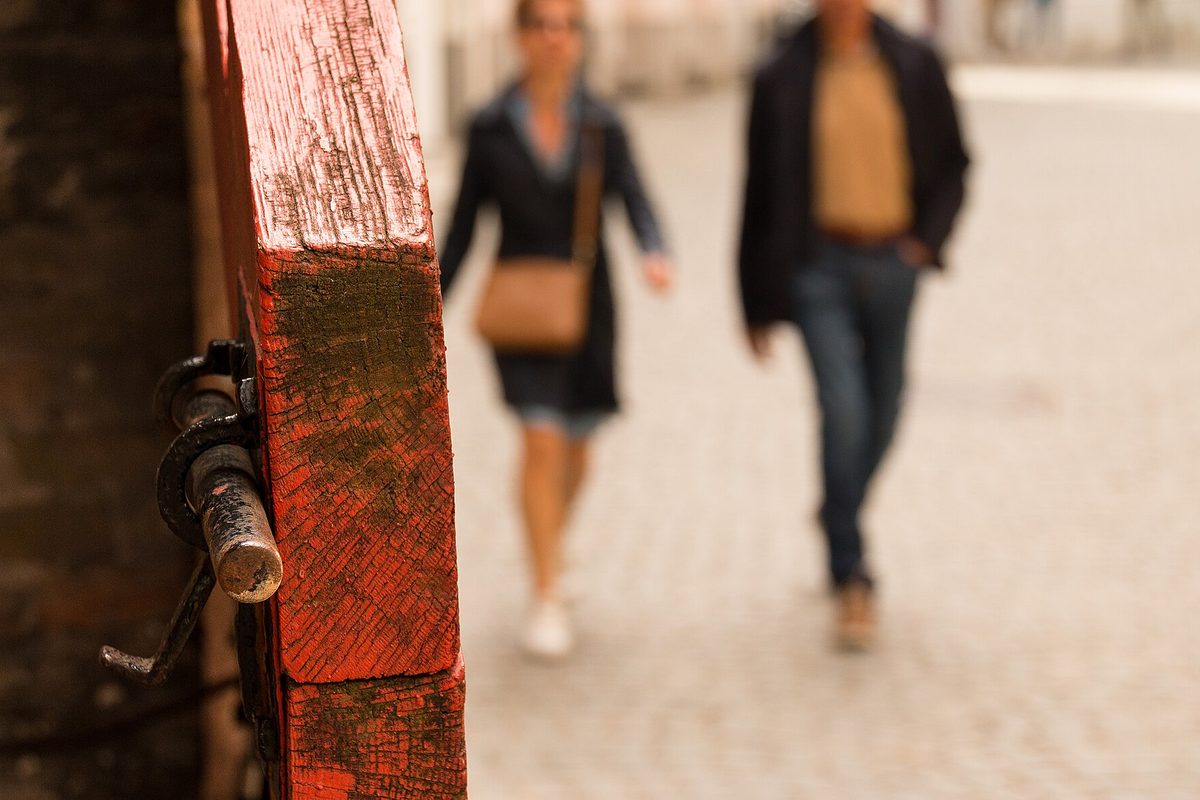 A black iron sliding bolt latch on the edge of a weathered red-painted wooden gate, with two people walking on a cobbled street blurred behind.