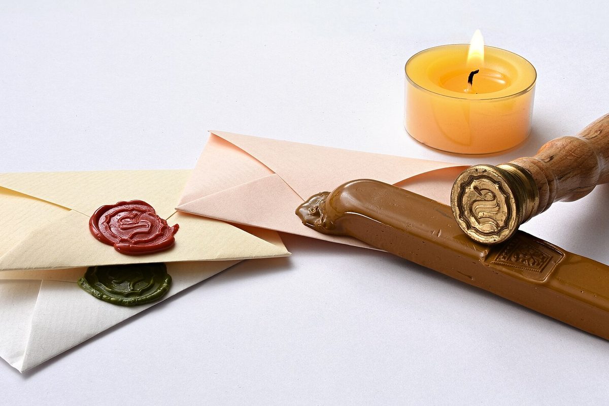 Two envelopes closed with red and green wax seals, next to a brass seal stamp, a stick of tan sealing wax and a lit tealight.