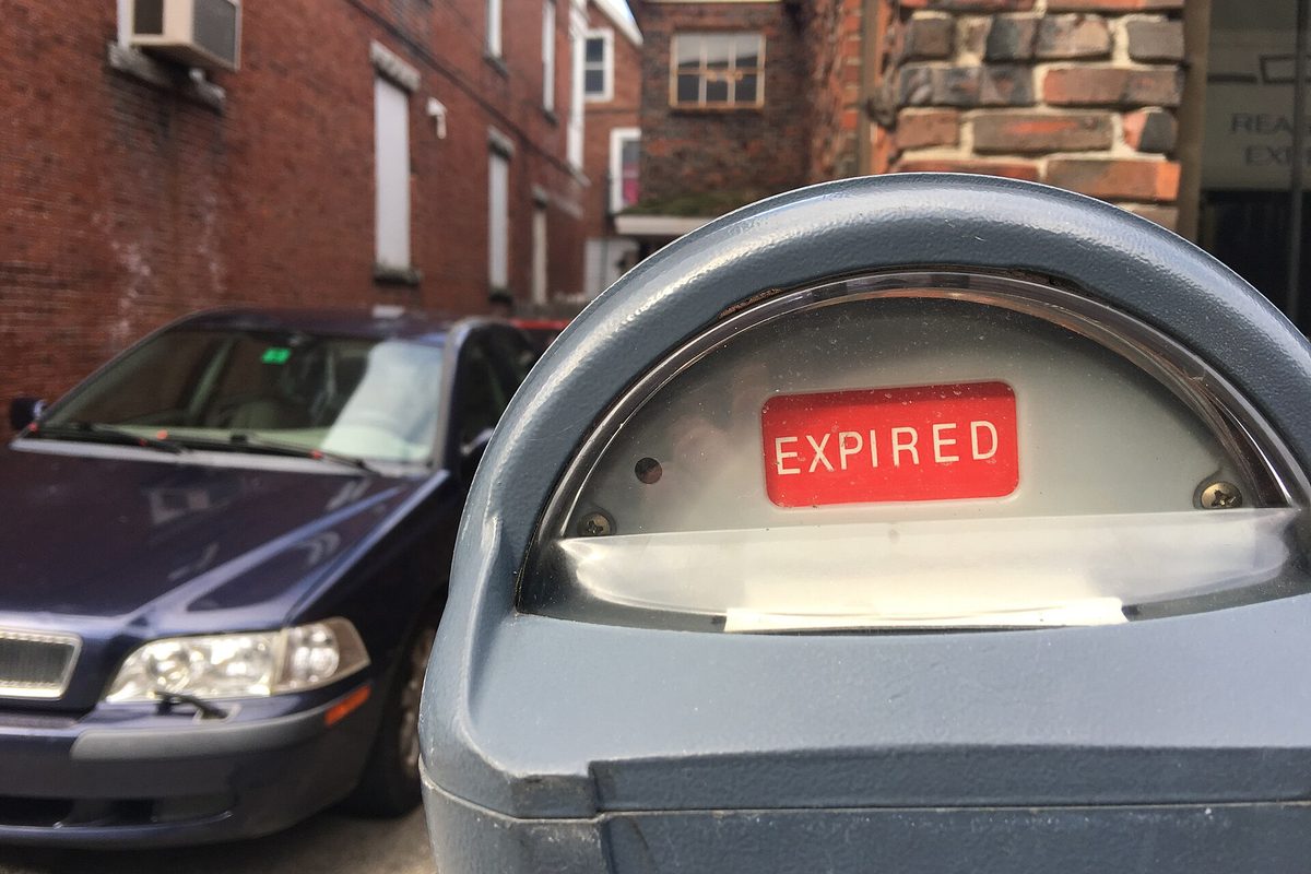 A grey metal parking meter showing a red EXPIRED flag behind its glass window, with a dark blue car and brick building behind.