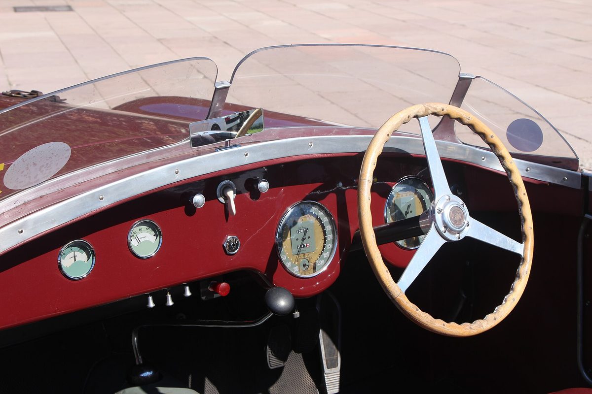 The maroon dashboard of a vintage open roadster with round chrome gauges, a wood-rimmed steering wheel and twin split windscreens.