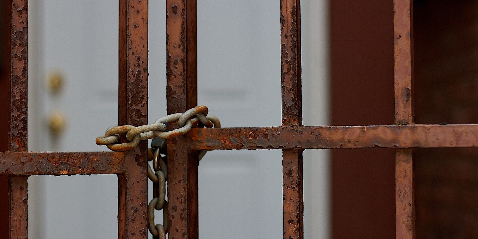 A galvanized steel chain and padlock wound around the rusted iron bars of a window grille, with a pale door blurred behind.
