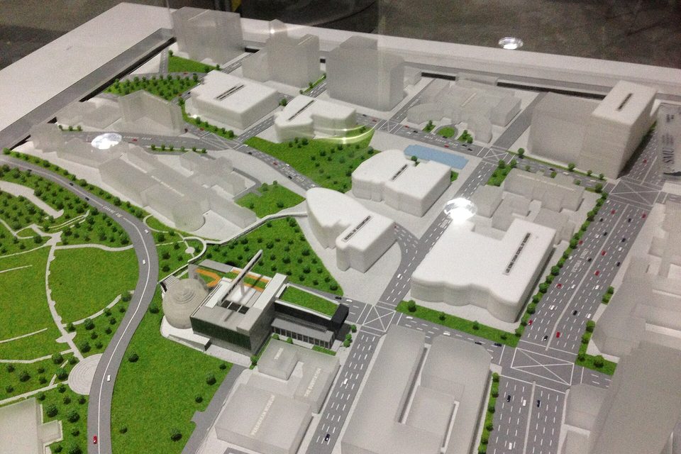 White scale model of a city district with roads, green parkland, trees and tiny cars on a table