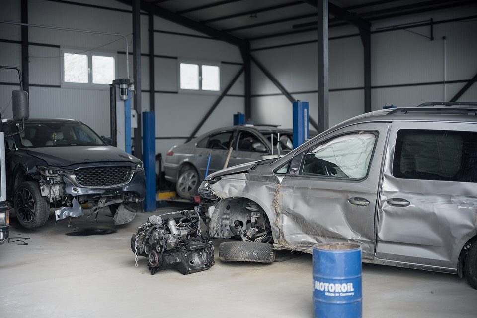 Body shop interior with wrecked vehicles, a bare engine on the floor and a blue oil drum