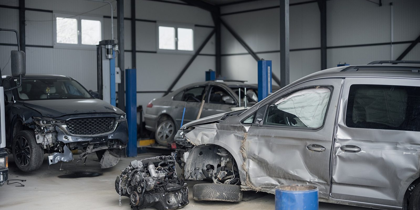 Collision repair bay holding several crash damaged cars beside blue lifts and a removed engine