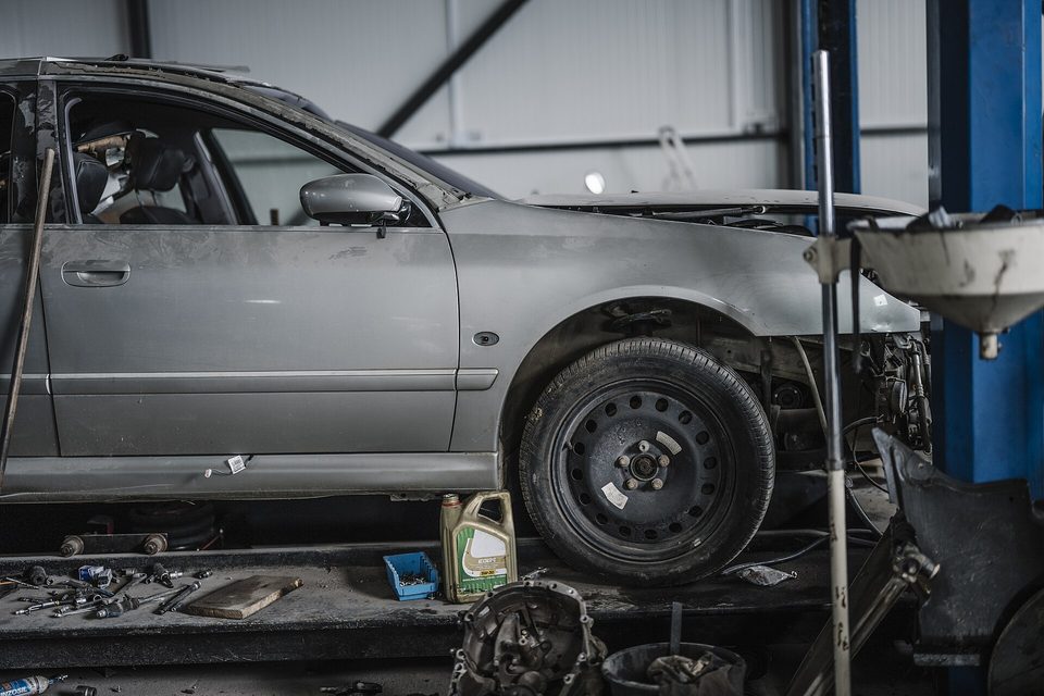 A partly stripped grey sedan on a two post lift with parts and an oil bottle on the floor