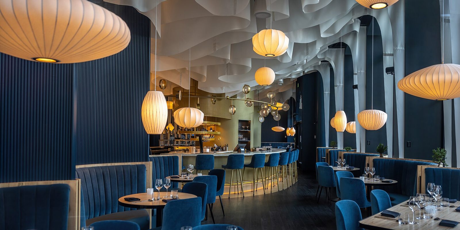 A modern restaurant dining room with blue velvet seating, a bar counter and rounded pendant lamps