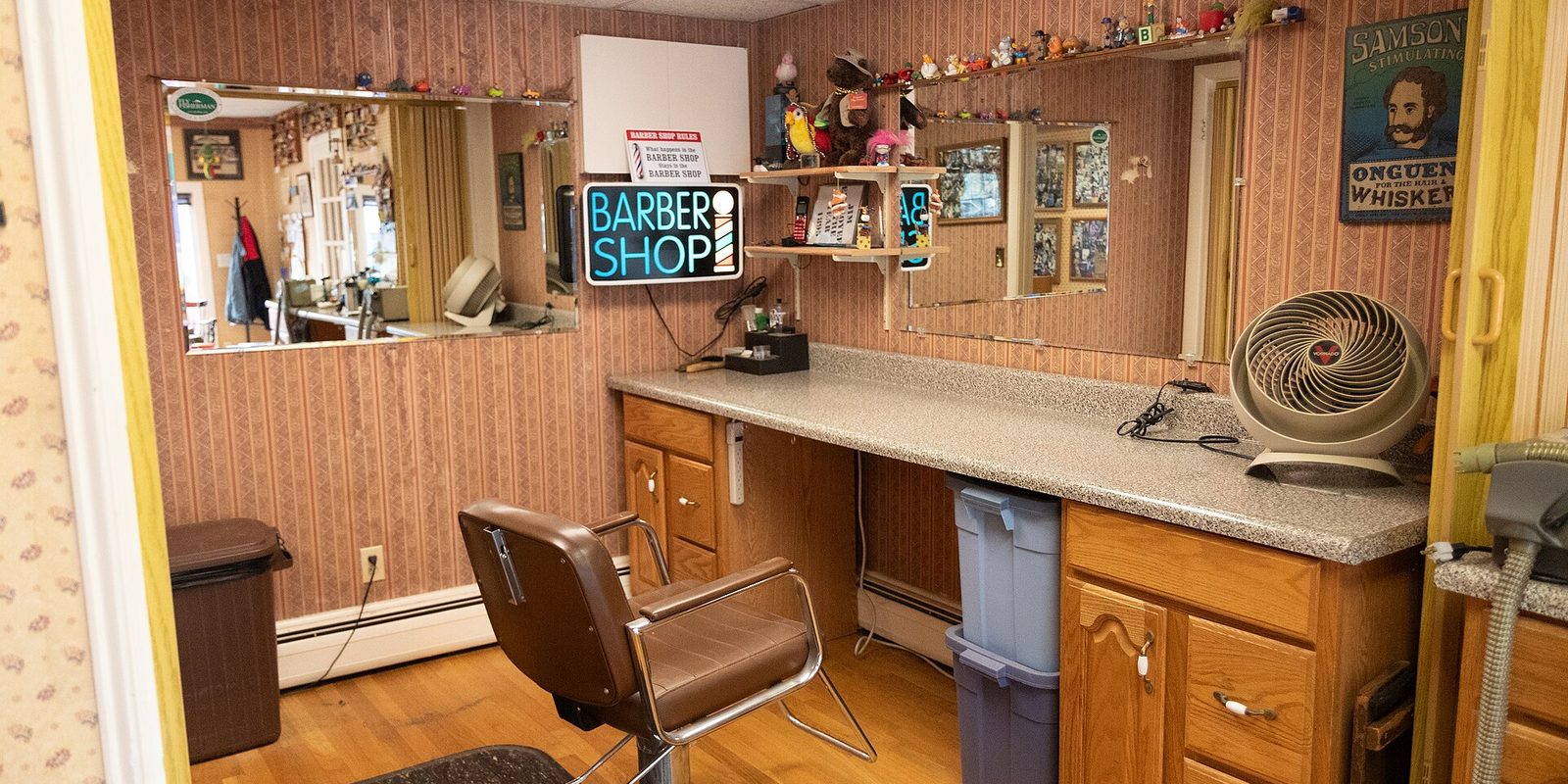 A small barber shop room with one styling chair, a long counter, wall mirrors and a lit Barber Shop sign