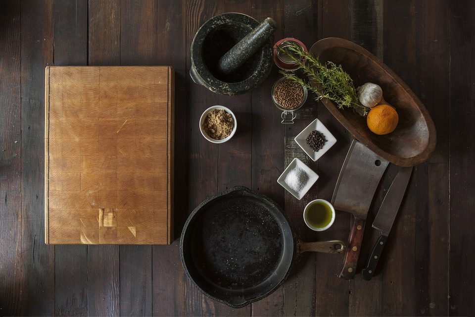 Overhead view of a dark wood table with a chopping block, skillet, mortar, knives, herbs and spices