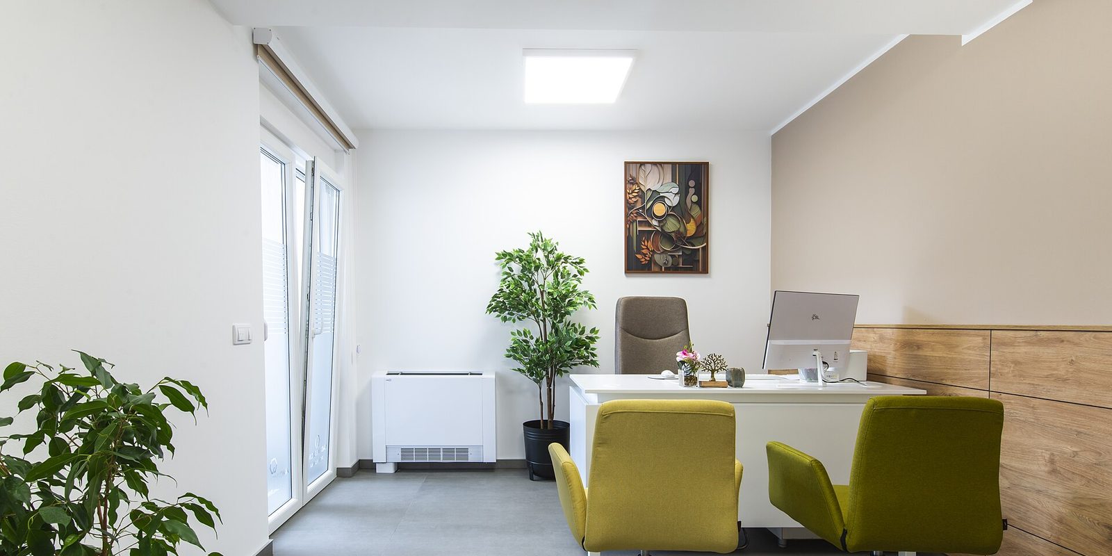 A small empty office with a white desk, monitor, green chairs, potted plants and wood cabinets