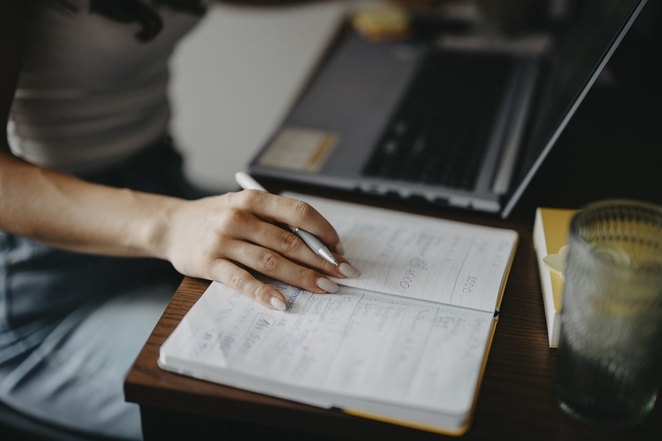 A hand holding a pen over a handwritten notebook beside an open laptop and a glass of water