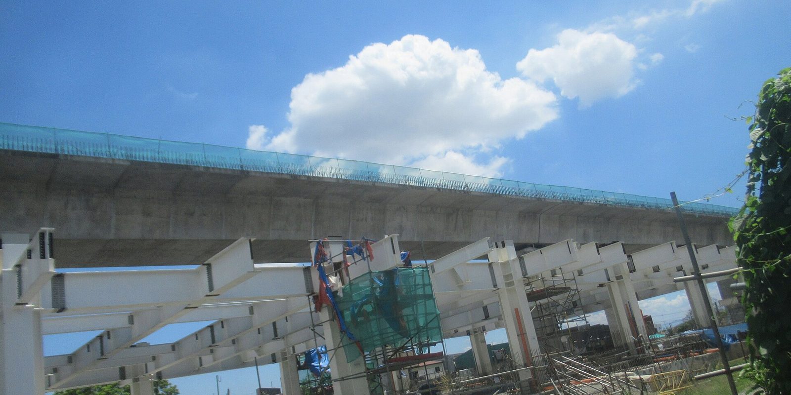 A concrete elevated roadway under construction above white steel beams, scaffolding and safety netting