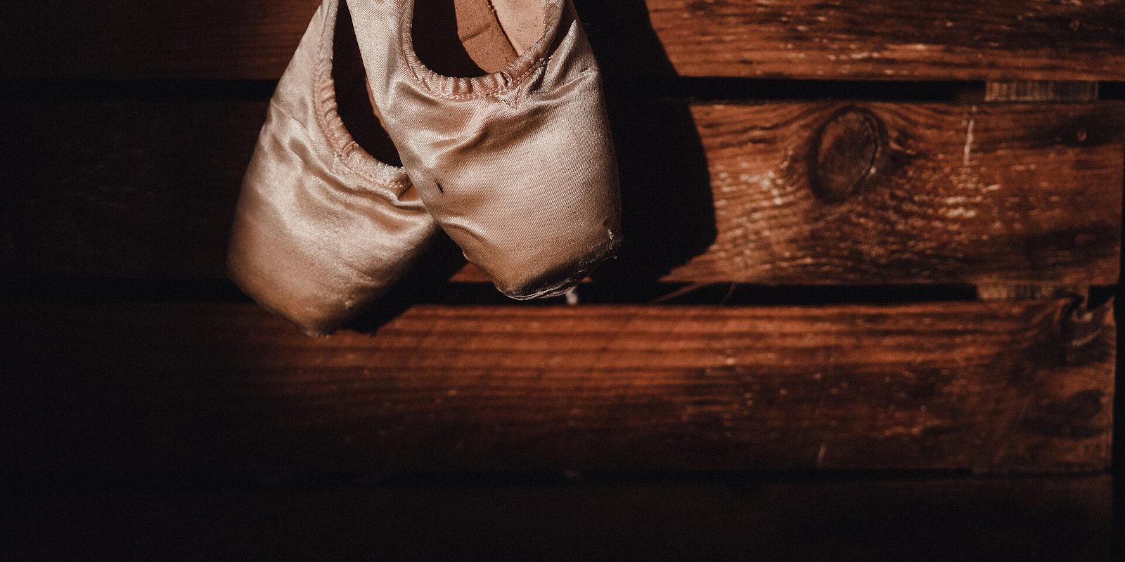 A pair of worn satin pointe shoes hanging against dark wooden boards in dim light