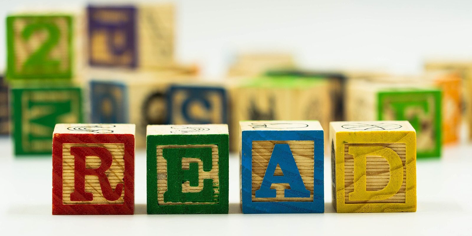 Wooden alphabet blocks spelling READ in red, green, blue and yellow, with more blocks blurred behind
