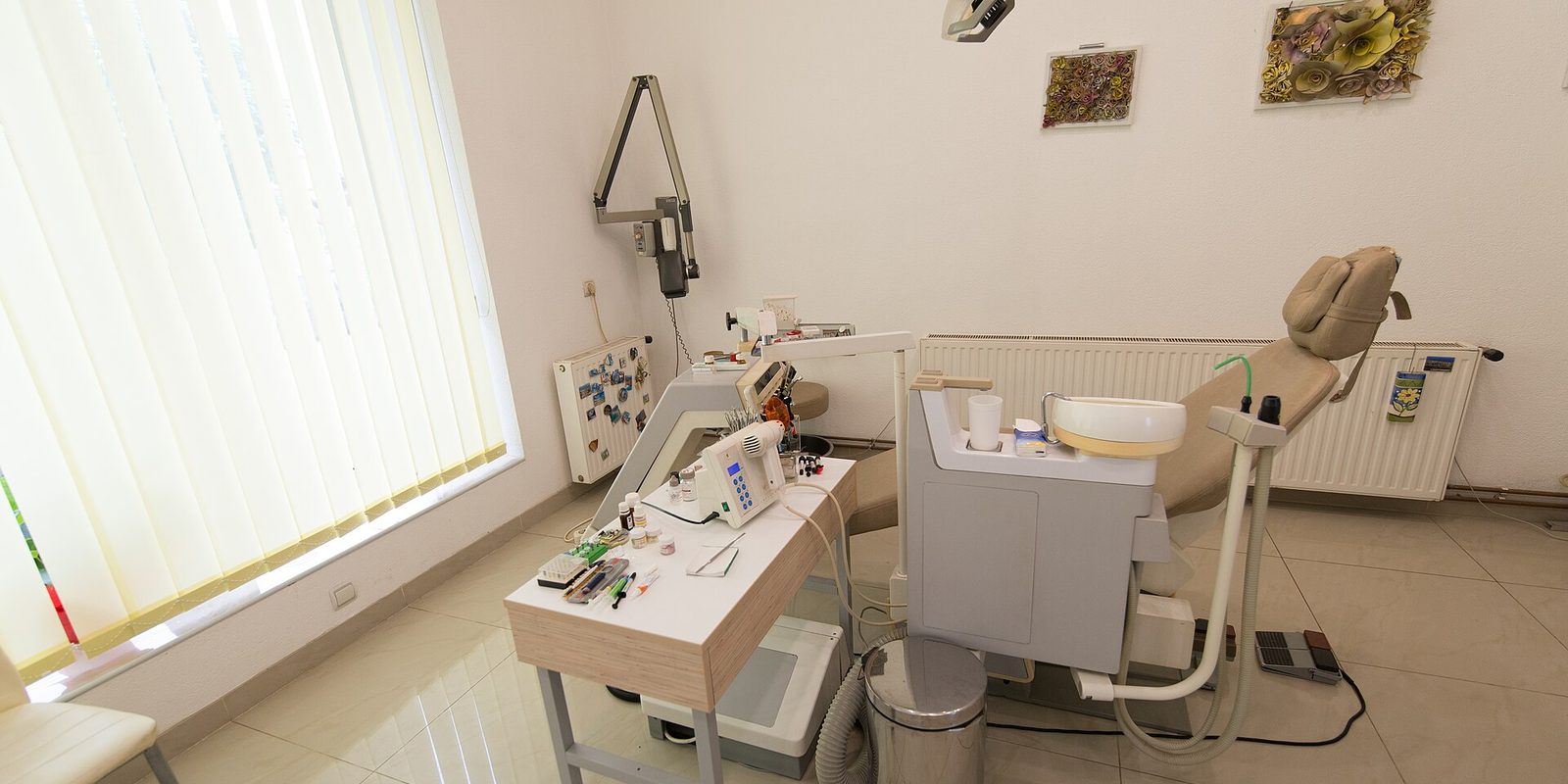 An empty dental treatment room with a reclining chair, instrument tray and bright window blinds