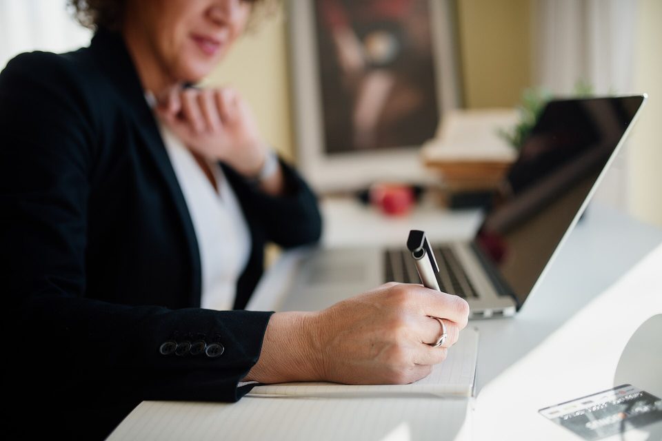 Someone in a dark blazer writing in a notebook at a desk with an open laptop behind it