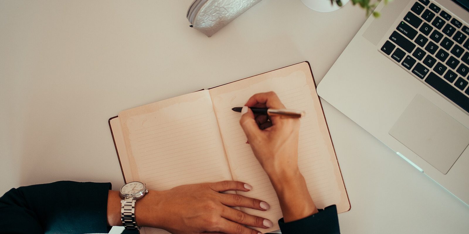 Overhead view of hands writing in a lined notebook on a white desk beside an open laptop