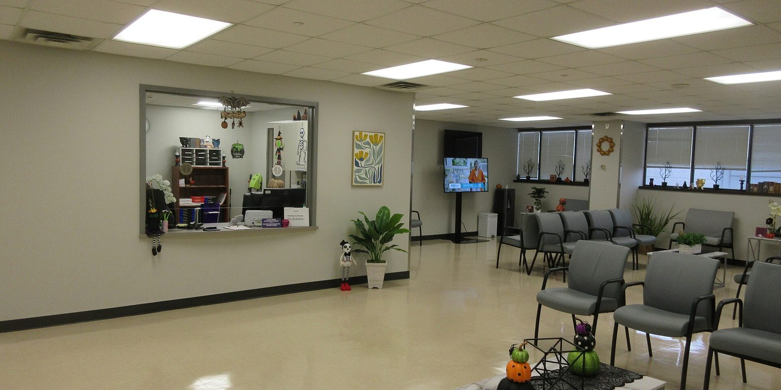 An empty medical waiting room with rows of grey chairs, a glass reception window and a wall-mounted television