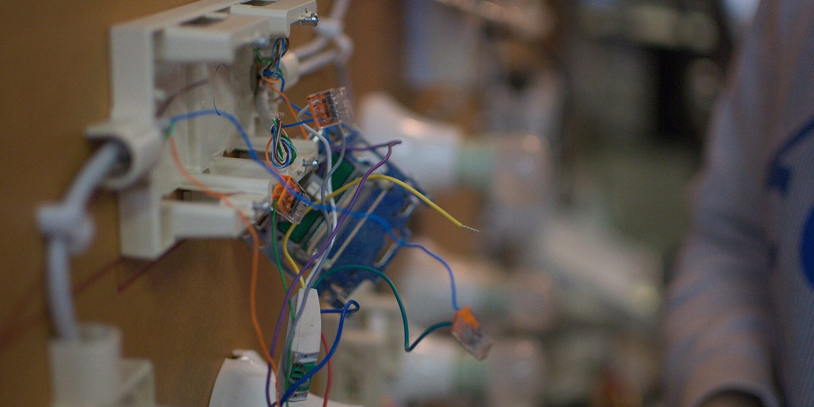 Close-up of a part-built wall switch box with coloured wires and lever connectors, workshop blurred behind