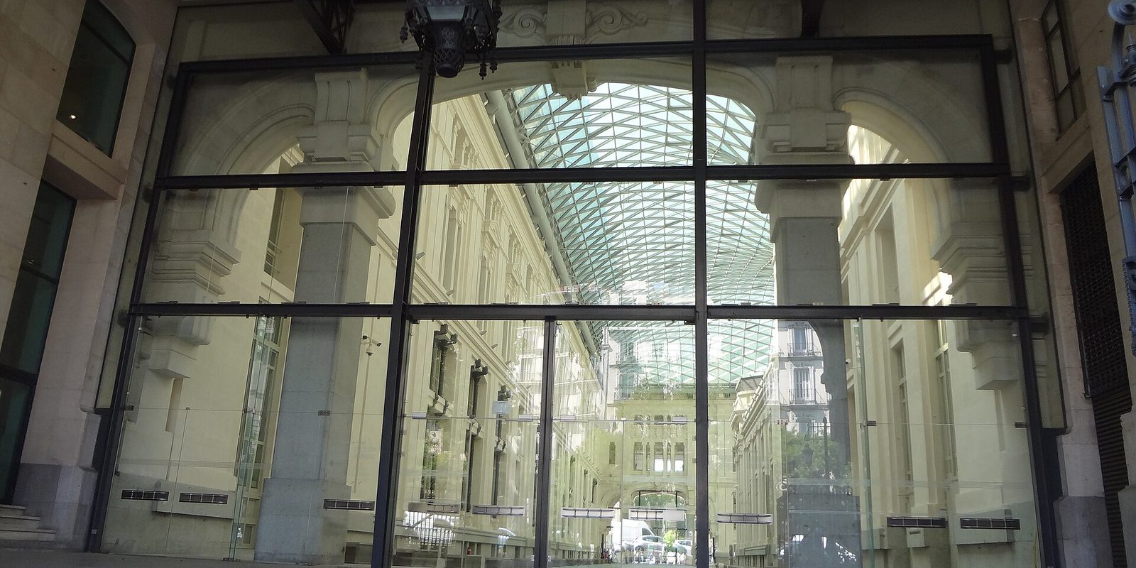 A tall glass entrance wall looking into a stone arcade covered by a curved glass and steel roof