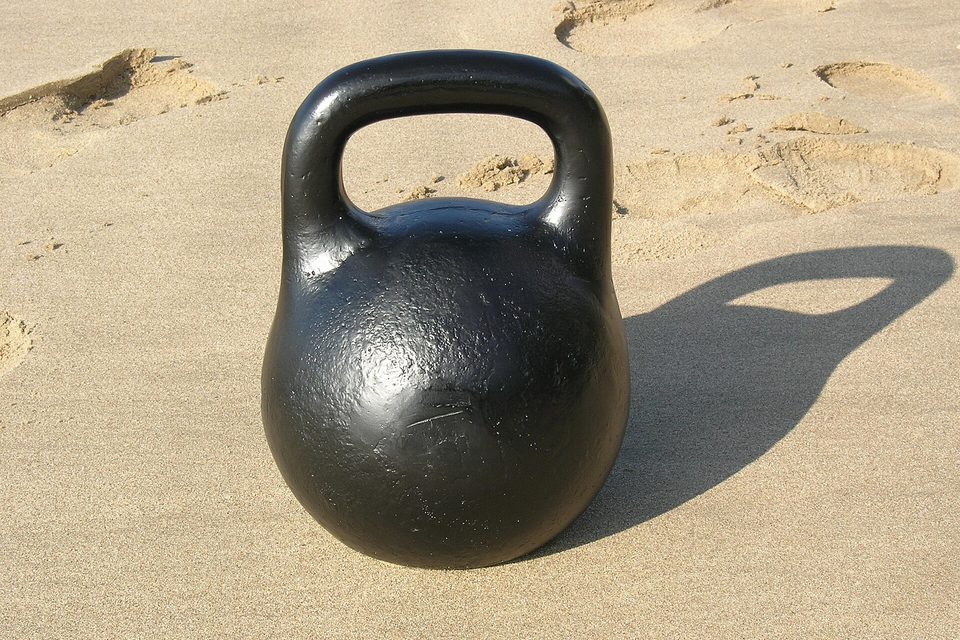 A black cast iron kettlebell resting on sand, casting a long shadow