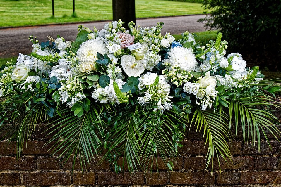 A long white and green floral arrangement with palm fronds resting on a brick wall outdoors
