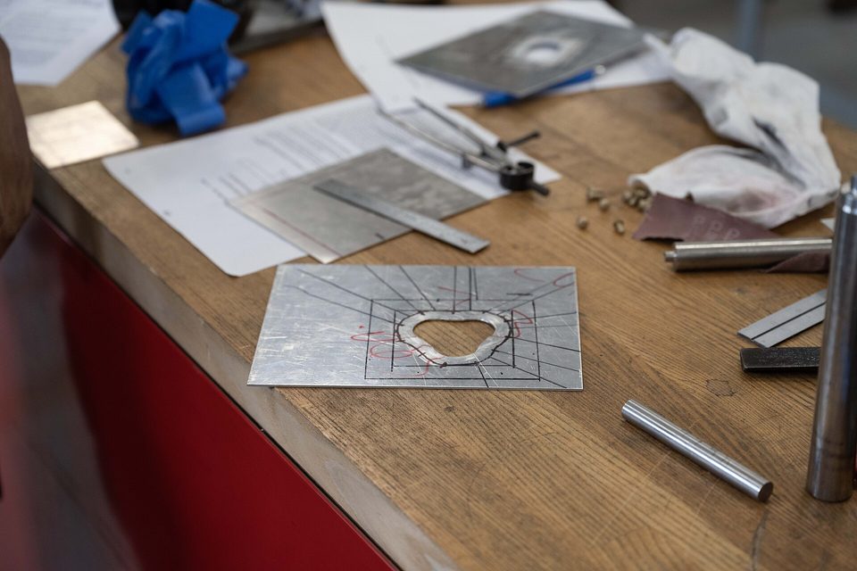 A workshop bench with a marked sheet metal plate, ruler, files and steel rod among paperwork