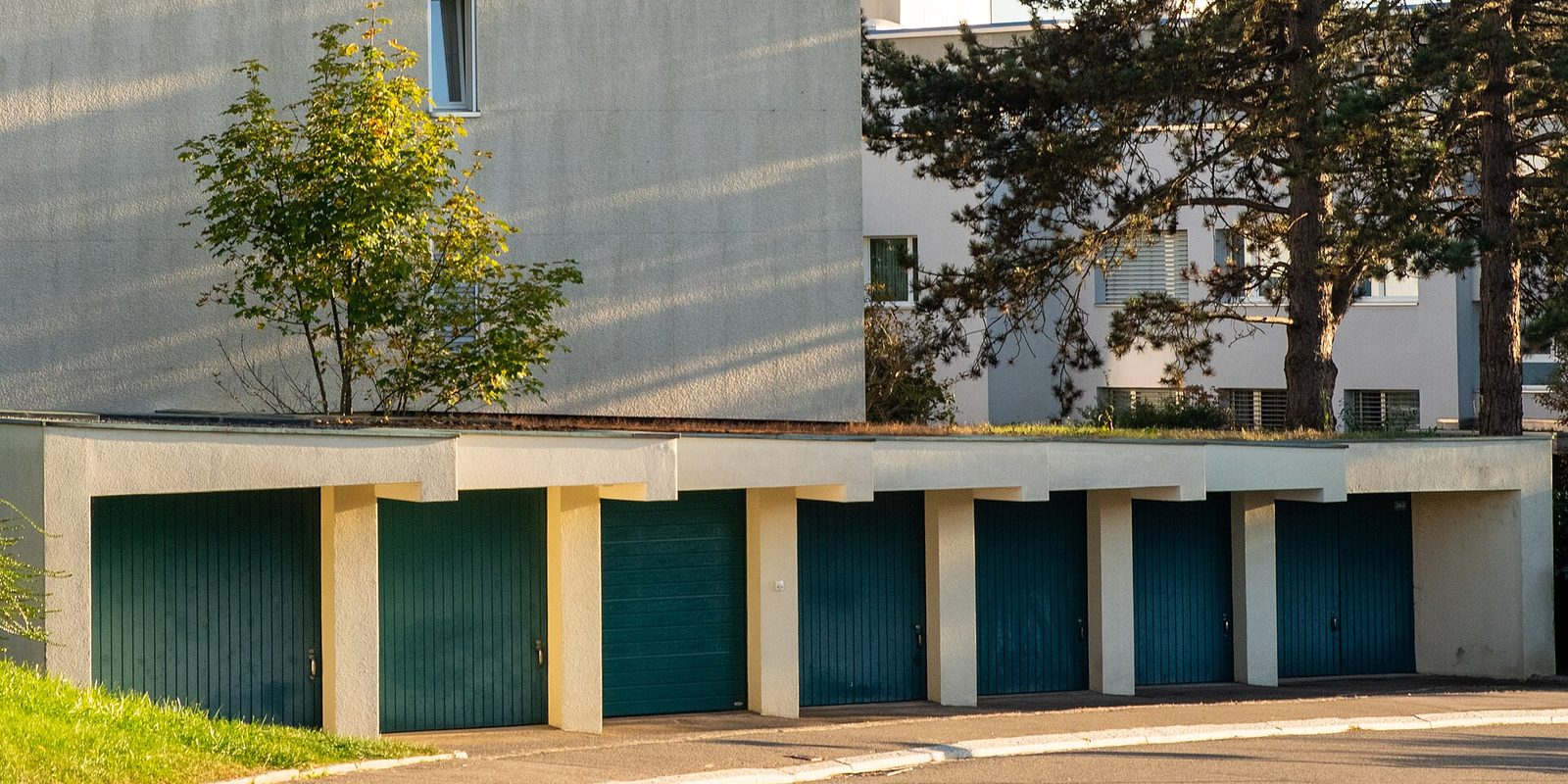 A row of concrete garages with dark green wooden doors below an apartment block in evening sun