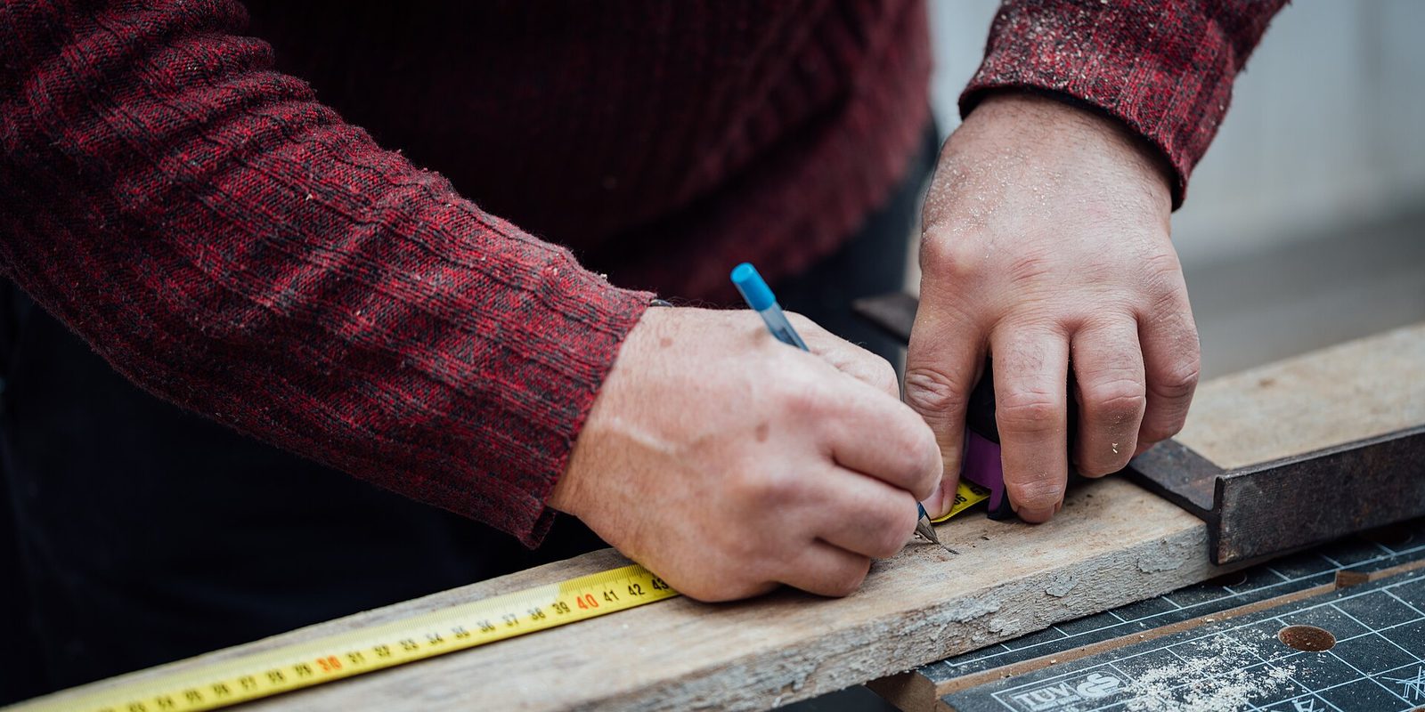 Close view of hands marking a plank with a pencil beside a yellow tape measure