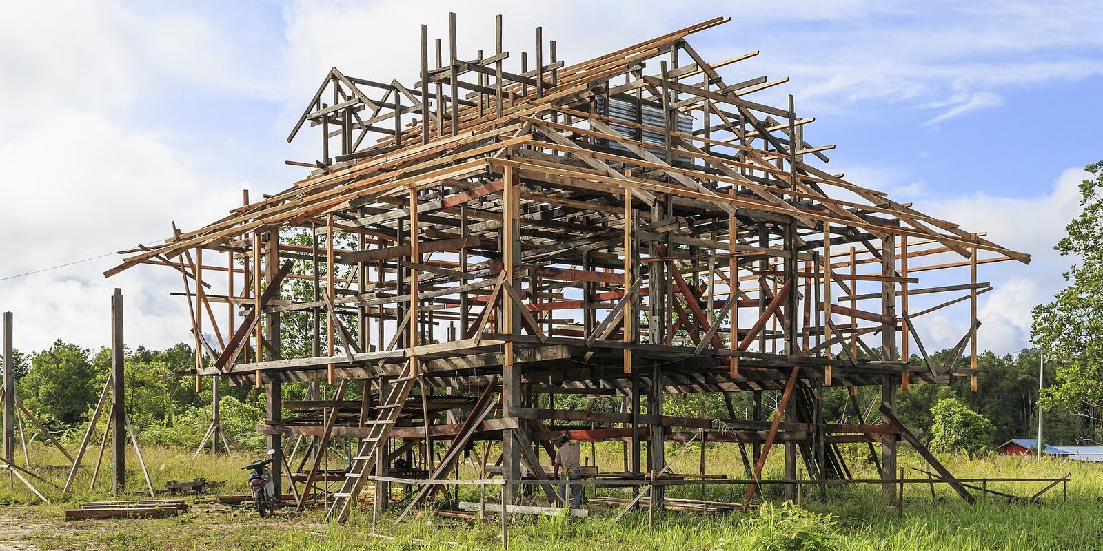 A large timber house frame standing in a grassy clearing under a blue sky, with one worker below
