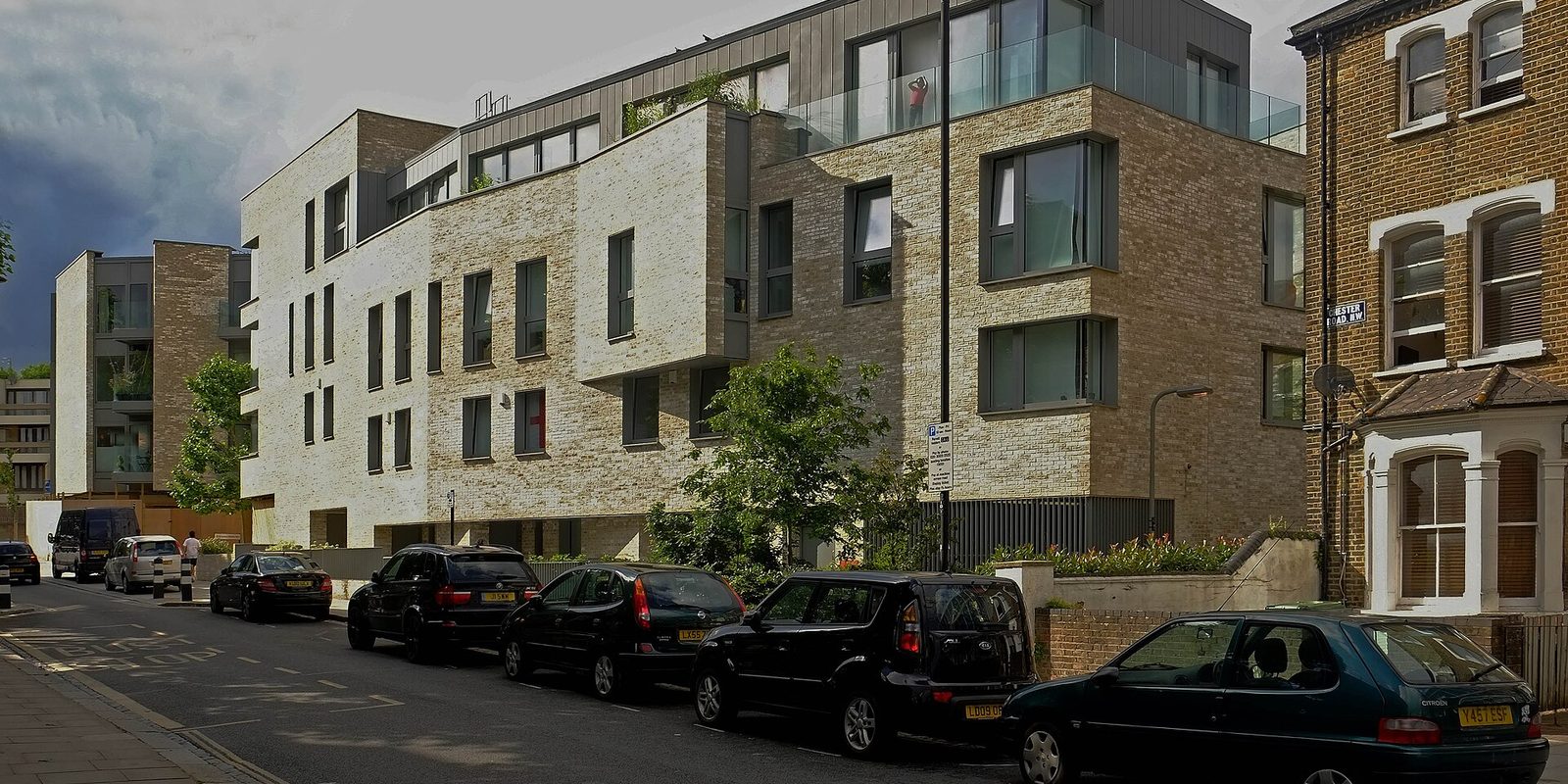 A modern brick apartment block on a residential street with cars parked along the kerb