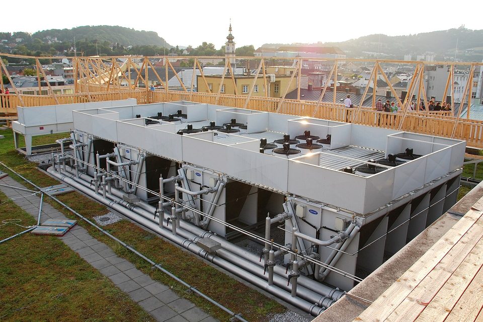Rows of large rooftop chiller units and grey pipework on a building roof above a town at dusk