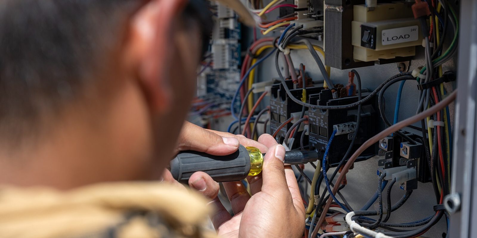 Close view over a technician's shoulder as they probe wiring inside an open HVAC control panel
