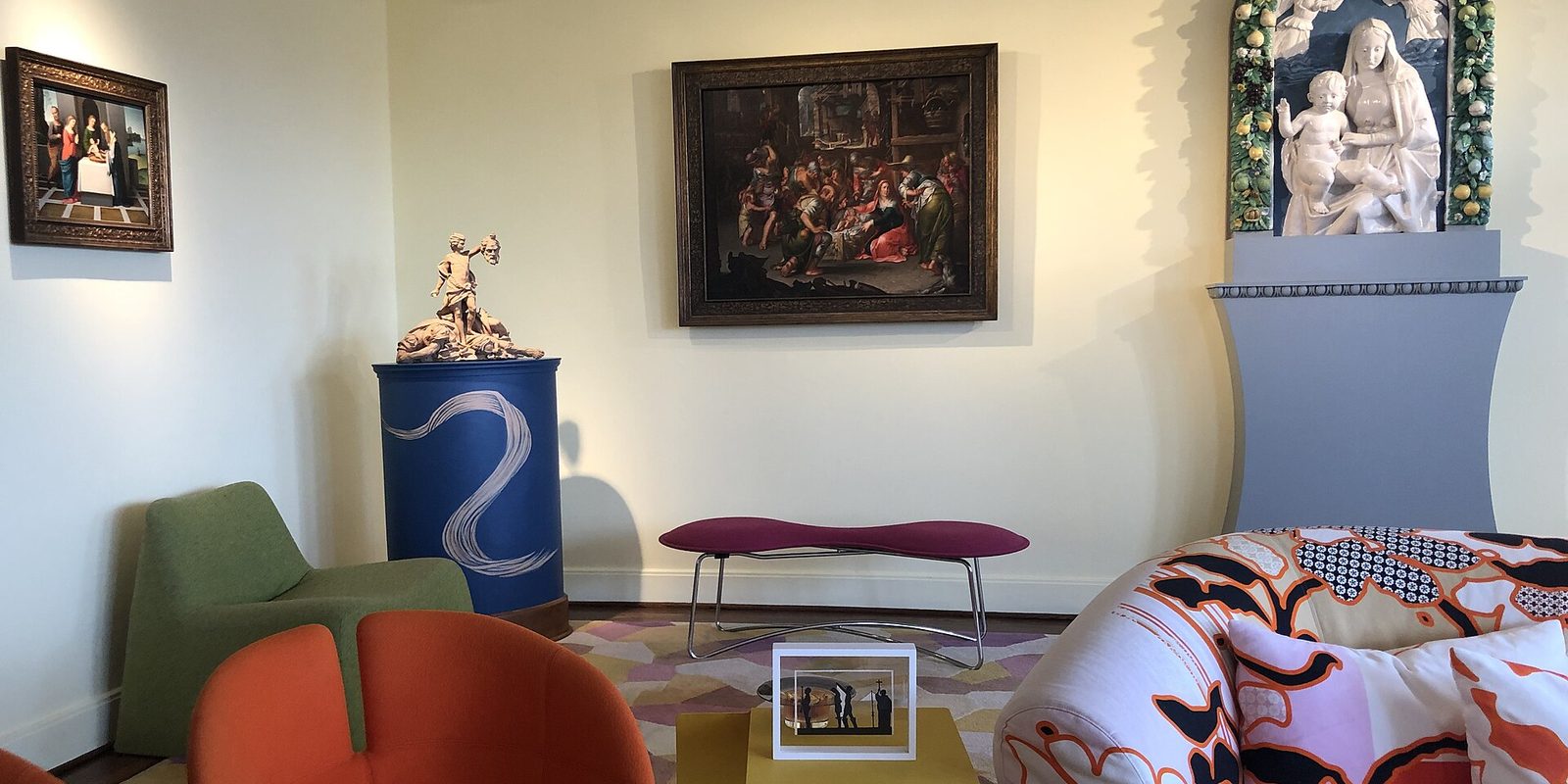 A cream sitting room with framed paintings, a green and orange armchair, pink bench and patterned sofa