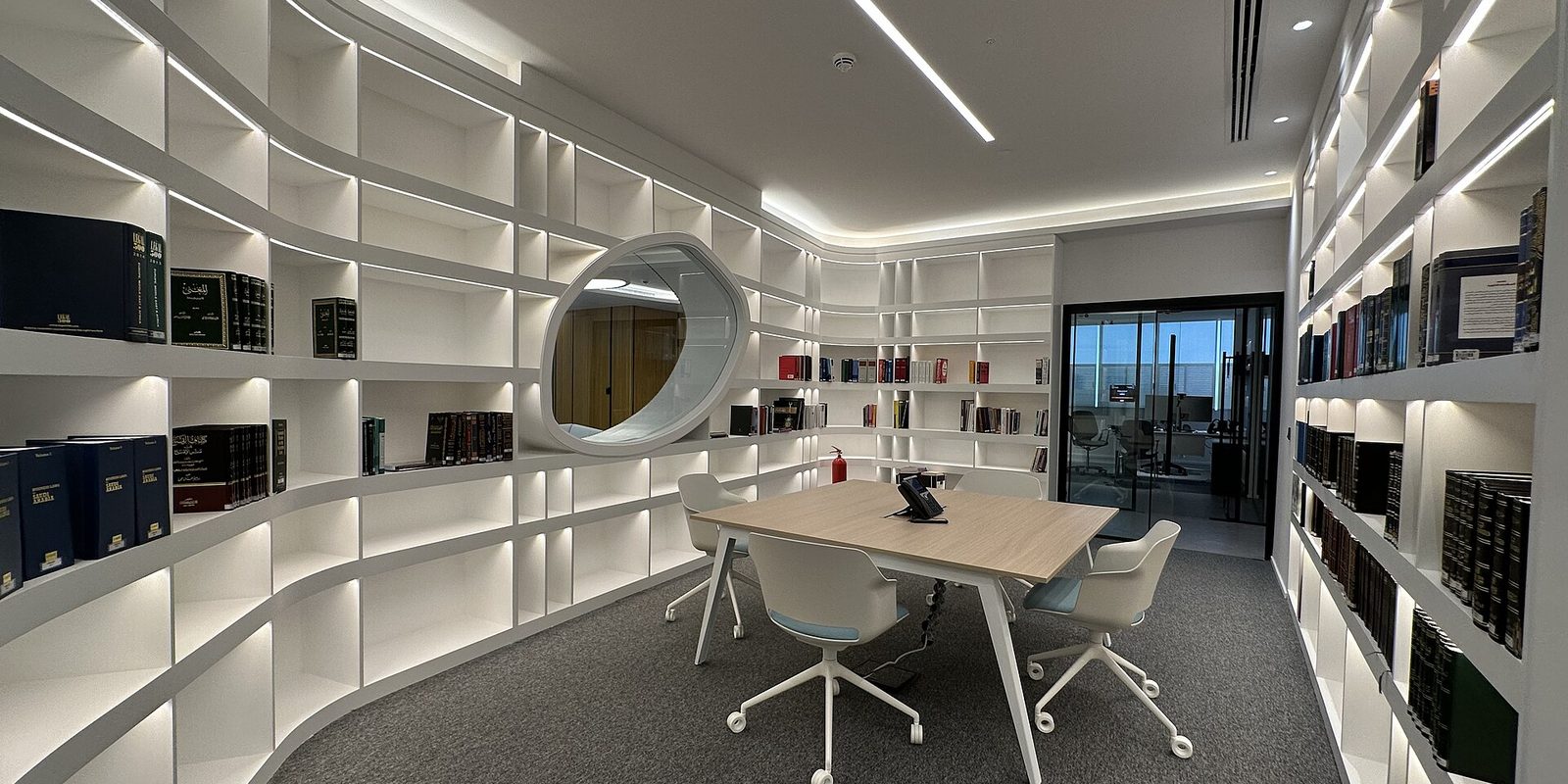 A brightly lit office library with curved white shelving, a small meeting table and four chairs